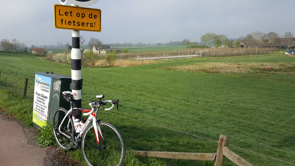 Wit-rode racefiets tegen een zwart-wit gestreepte paal, met open weiland erachter.