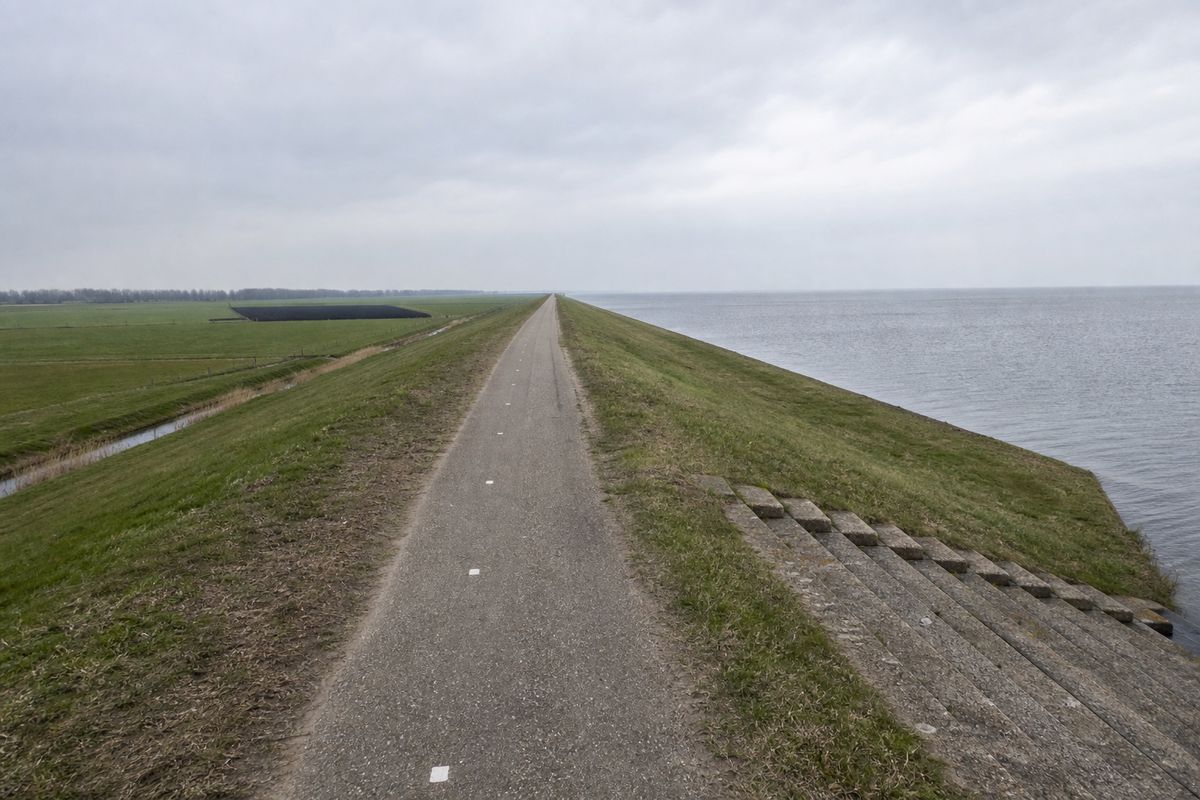 Fietspad op de kruin van een lange rechte zeedijk, met het IJsselmeer aan de ene en laag polderland aan de andere kant.